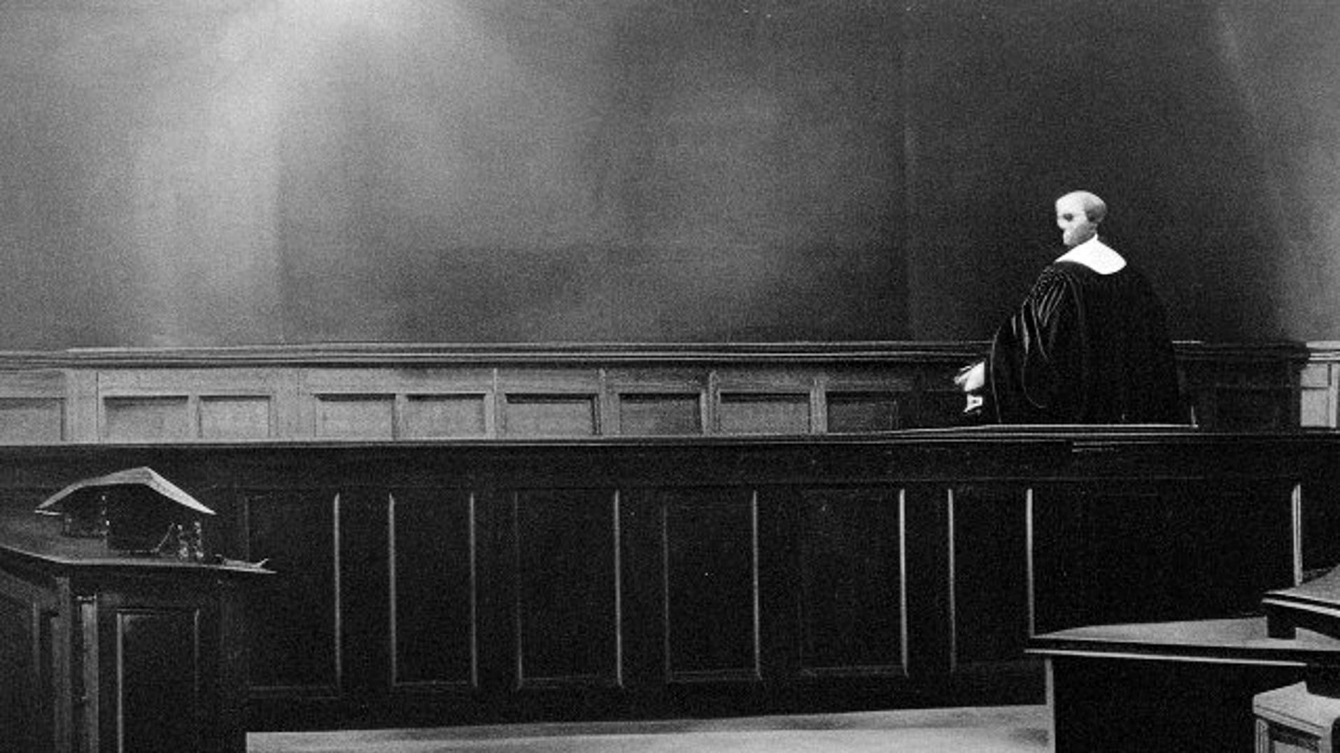 A solitary figure stands in a dimly lit courtroom, their gaze piercing the emptiness. The stark black-and-white contrast underscores the weight of justice and the gravity of decisions yet to be made, evoking a sense of solitude in the pursuit of accountability.